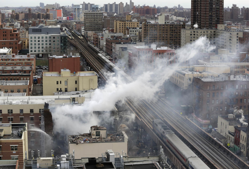 Firefighters, lower left, pour water on the site of a building explosion, as a Metro-North commuter train, right, passes the site on Thursday, March 13, 2014 in New York. Rescuers working amid gusty winds, cold temperatures and billowing smoke pulled four additional bodies Thursday from the rubble of two New York City apartment buildings, raising the death toll to at least seven from a gas leak-triggered explosion that reduced the area to a pile of smashed bricks, splinters and mangled metal. The explosion Wednesday morning in Manhattan's East Harlem injured more than 60 people. (AP Photo/Mark Lennihan)