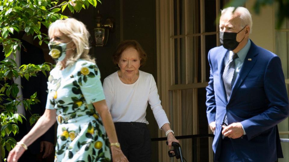 rosalynn carter uses a walker as she moves next to a window, two people walk in the forefront of the photo frame