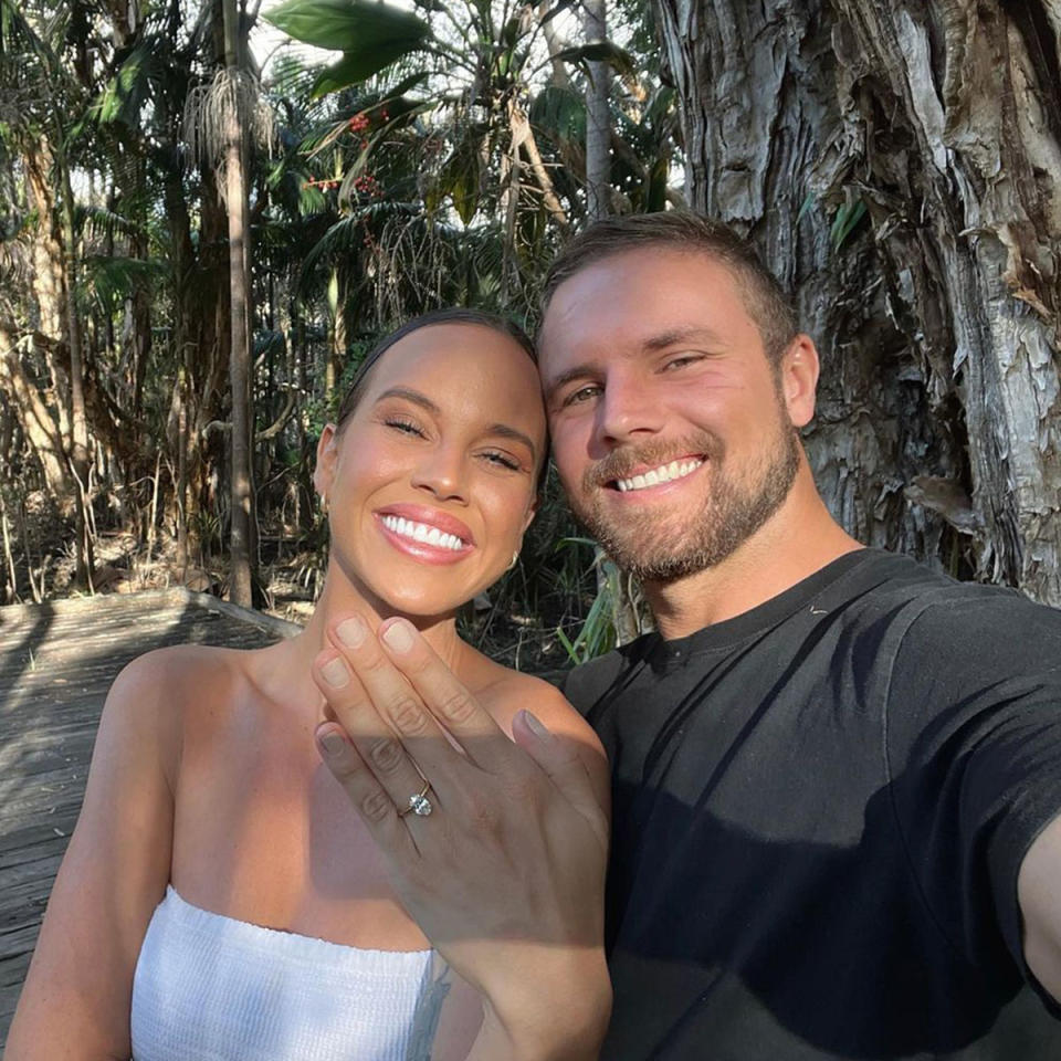 Bonnie Anderson and Sam Morrison are engaged smile for the camera while Bonnie shows off her diamond ring. Sam is in a black t shirt, Bonnie is in white.