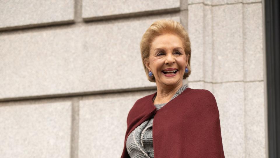 carolina herrera stands outside a stone building and holds a handrail, she smiles and looks right of the camera, she wears a gray plaid skirt suit and a burgundy cape with blue earrings