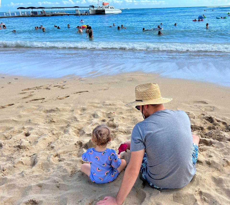 A child and her father wearing a straw hat sitting on the beach.