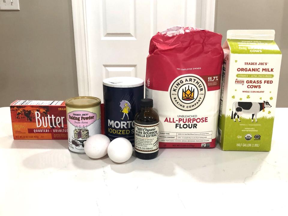 Containers of butter, baking powder, salt, vanilla extract, flour, and organic milk on a white counter. There are also two eggs.