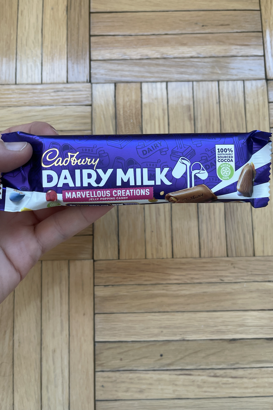 Hand holding a Cadbury Dairy Milk Marvellous Creations chocolate bar over a wooden floor