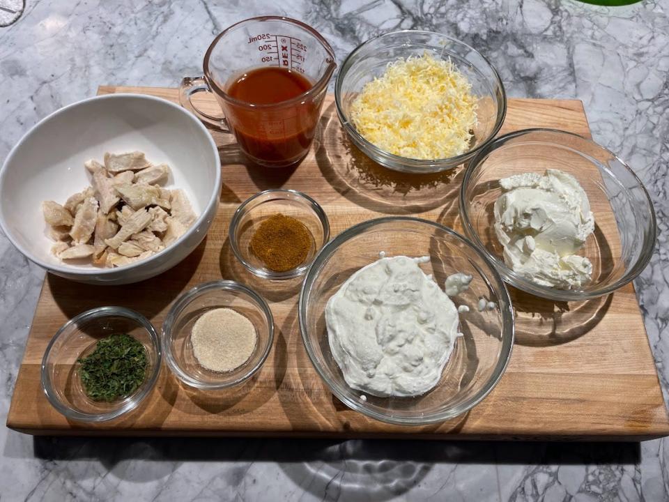 Cutting board with bowls of ingredients including sour cream, shredded cheese, seasonings, a liquid measuring cup filled with hot sauce, and canned chicken