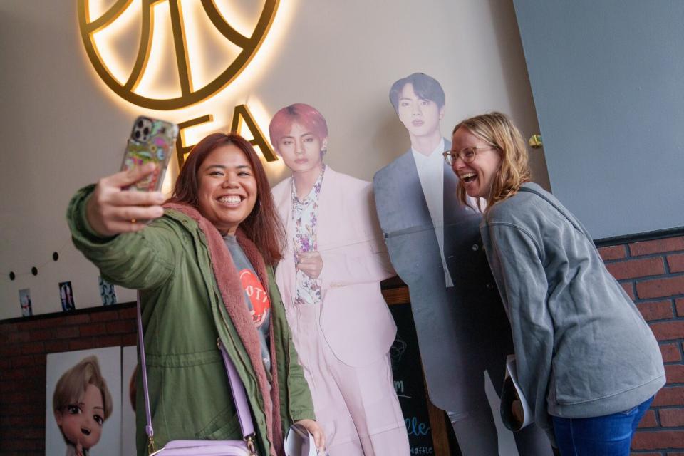 Two women take selfies with a pair of life-size K-pop artist standees.