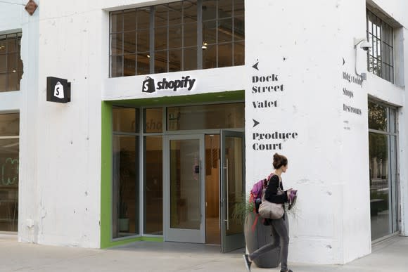 A young woman walks by the entrance of Shopify LA. The store is in a white concrete building and the company logo is above the door.