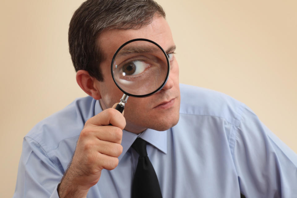 A man in a blue dress shirt and black tie looks through a magnifying glass, enlarging his eye