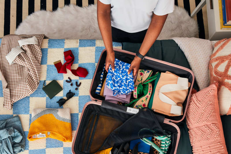 overhead shot of a person folding a floral shirt in suitcase that's on a bed with blue and white checkered sheets with other clothes and accessories surrounding the suitcase