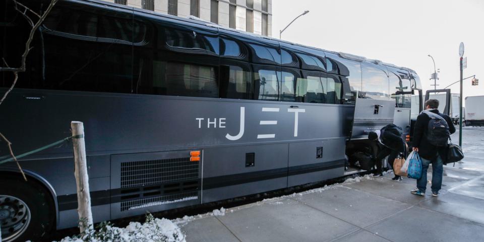 A matte black bus that reads "The Jet" on the side. Passengers with bags are boarding the bus or putting their bags away into the lower storage compartment.