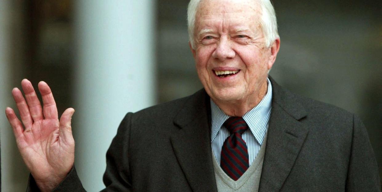 jimmy carter smiles and waves to the camera, he is wearing a blue and white striped collared shirt with a navy and dark red diagonal striped tie, a light gray v neck sweater is on top of the shirt and tie, and he also wears a dark gray suit jacket