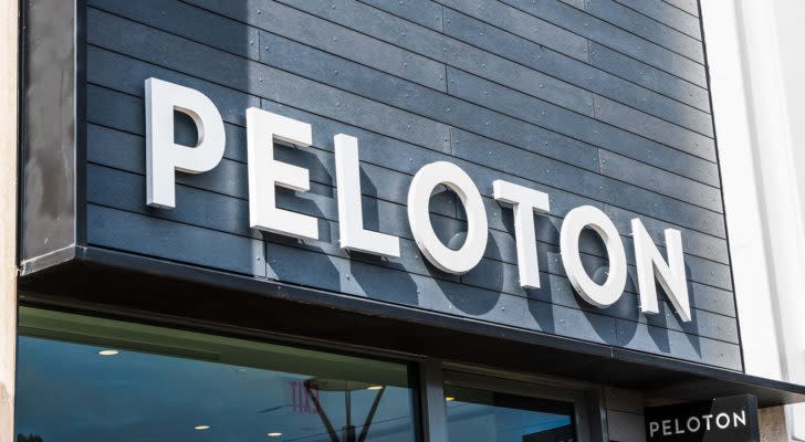 An image of a grey store front with a white "Peloton" logo on the building and a smaller black sign with a white "Peloton" logo on it in front of a glass door and window.