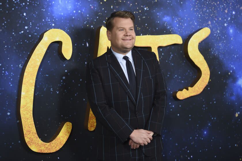 A man in a suit and tie stands in front of a "Cats" movie backdrop and clasps his hands in fig-leaf position