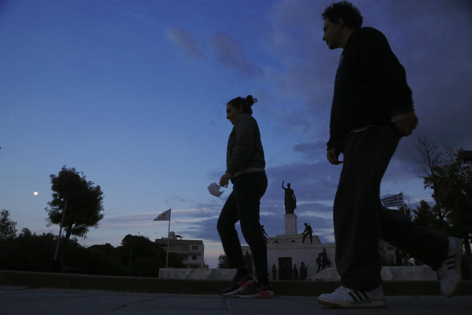 A woman holds a face mask and a man walk in front of the Liberty monument in central capital Nicosia, Cyprus, Wednesday, Jan. 27, 2021. Cyprus' health minister Constantinos Ioannou said that the first to re-open as of Feb. 1st will be hair and beauty salons followed a week later by retail stores, shopping malls and elementary schools. Students in their final year of high school will also go back to classes on Feb. 8. (AP Photo/Petros Karadjias)