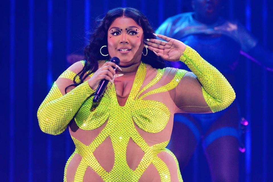 Lizzo speaking into a microphone while raising her hand to her ear and wearing a yellow figure-hugging dress.