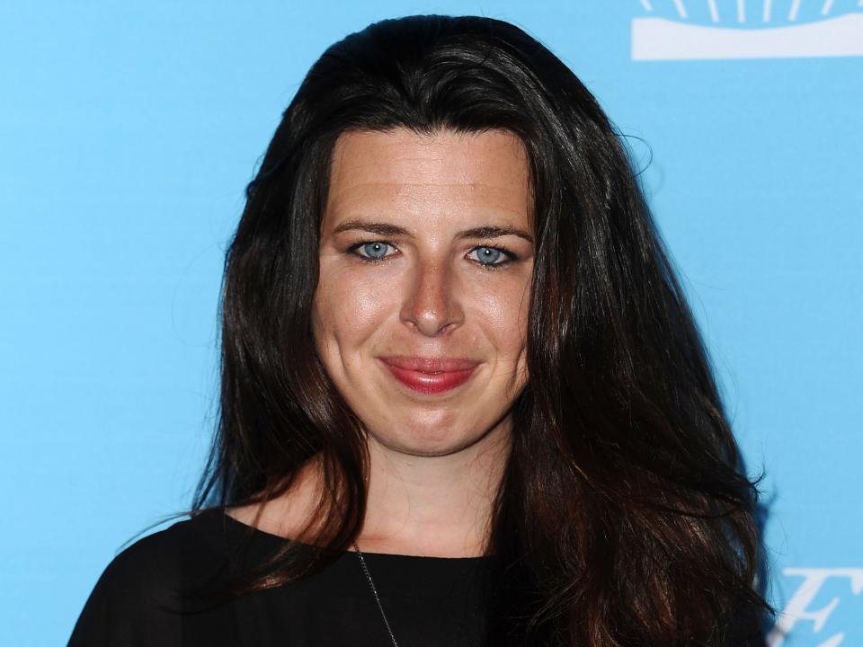 Heather Matarazzo smiling while wearing a black top and standing in front of a light blue background.