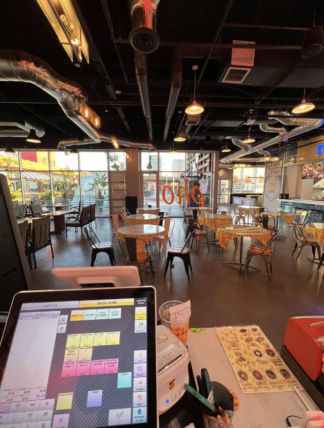 An empty, modern-style pizza restaurant seen from behind the order counter, with tables and chairs set up for dining