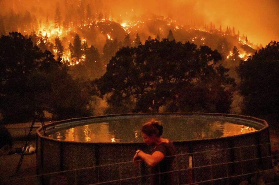 A woman leans on a fence next to an above-ground pool as wildfire burns on a nearby ridge.