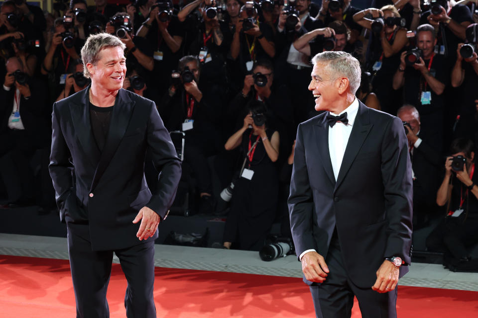 Brad Pitt and George Clooney smile on the red carpet with photographers in the background. Brad wears a black suit and George a black tuxedo