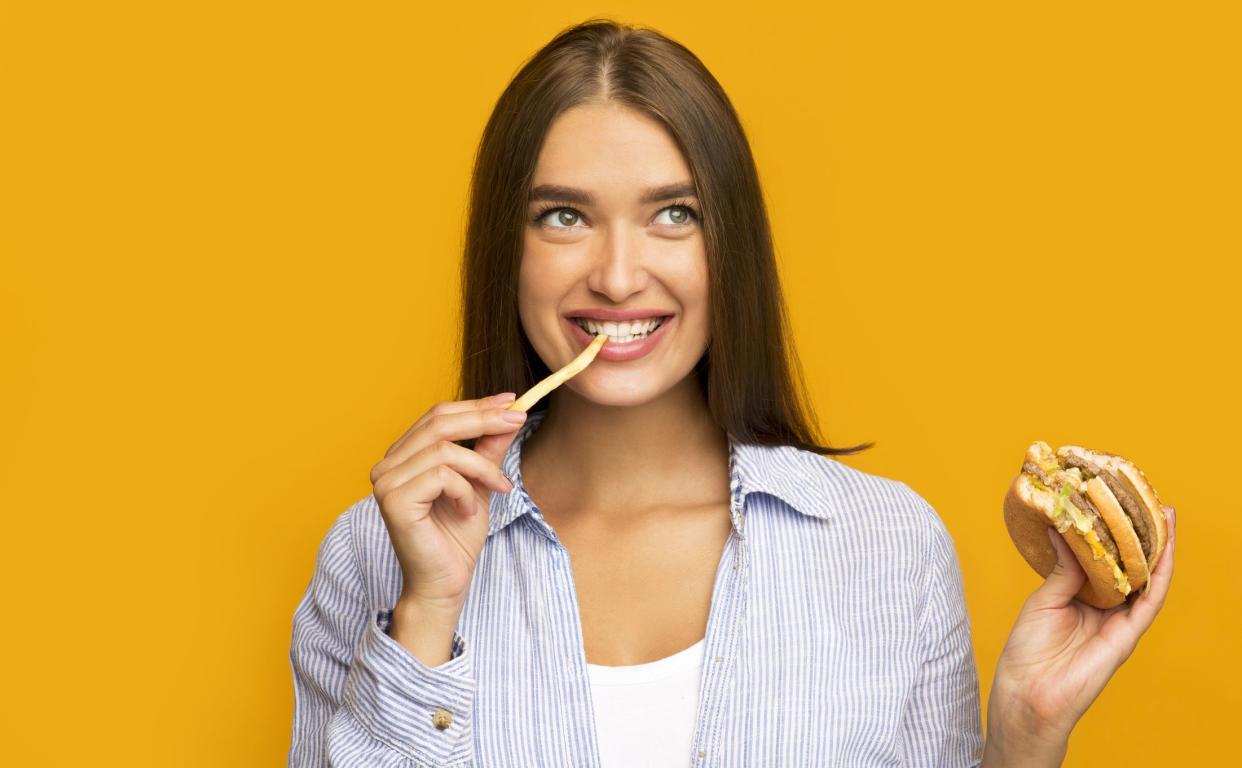 Unhealthy Food. Happy Girl Eating Burger And French Fries Standing Over Yellow Studio Background