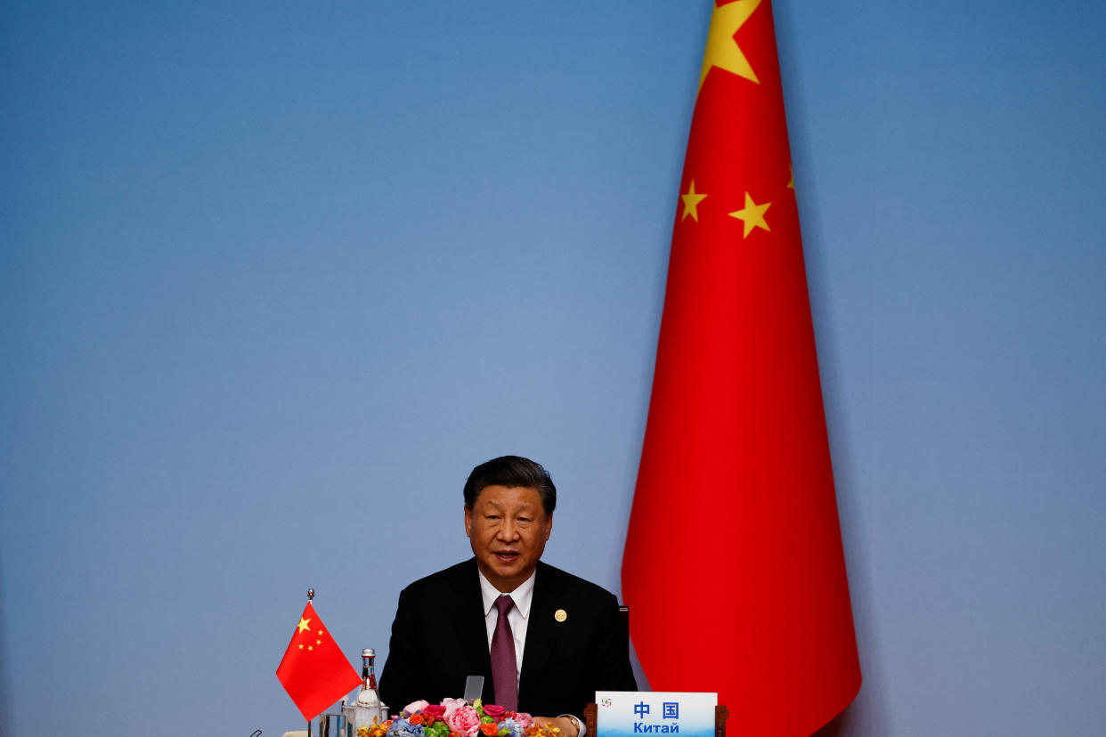 Chinese President Xi Jinping sits next to a Chinese flag against a blue backdrop at a desk with flowers, another flag, a bottle and a microphone.
