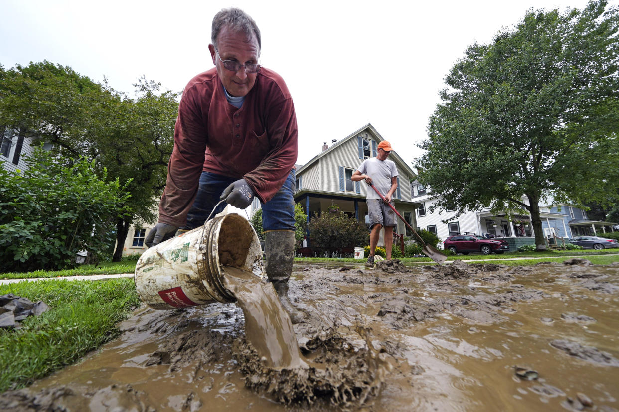 A man dumps a bucket of muddy water while another man clears away mud in front of a house.