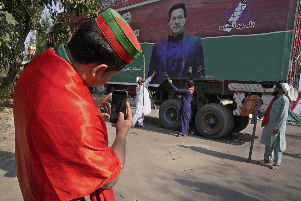 A supporter of former Prime Minister Imran Khan takes photos with mobile phone to his colleagues with huge Khan's poster painted on a truck neat the Khan's residence, in Lahore, Pakistan, Thursday, March 16, 2023. A Pakistani court on Thursday extended a pause for a day in an operation aimed at arresting the former premier Khan, a sign of easing tension in the country's cultural capital of Lahore where 24-hours-long clashes erupted this week when police tried to arrest Khan for failing to appear before a court in the capital. (AP Photo/K.M. Chaudary)