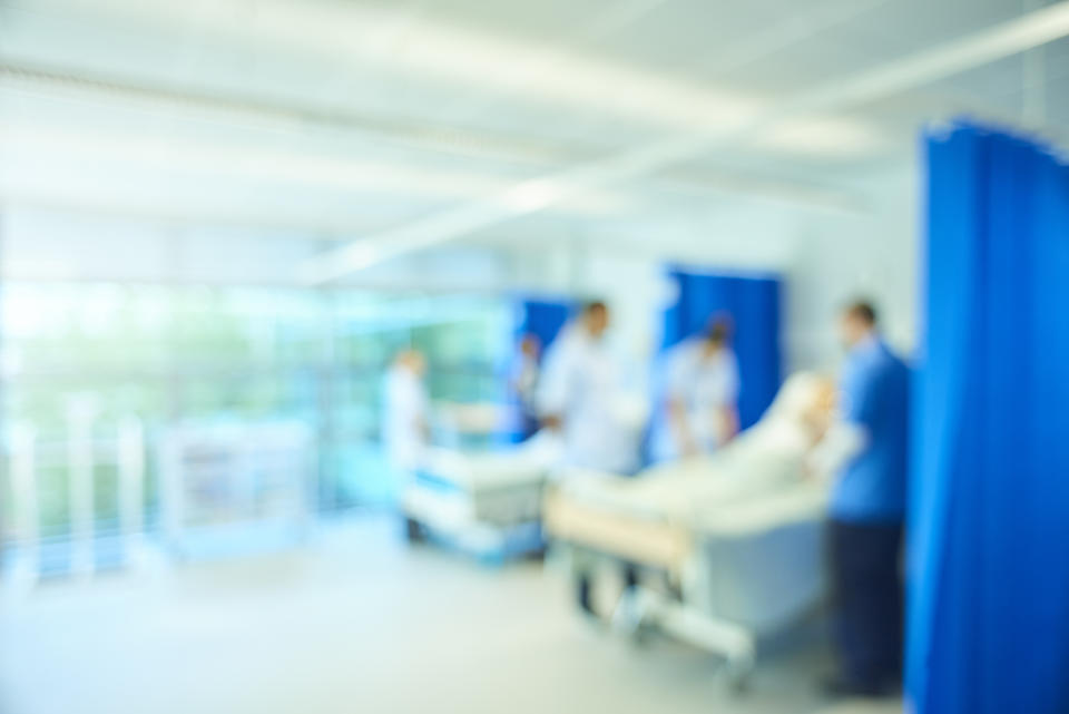 A  senior female nursing sister demonstrates the the various equipment on the training ward whilst a male staff nurse shows the medical mannequin to another group of medical student nurses . They are all standing around the hospital beds . The whole scene is defocussed to be used as a background