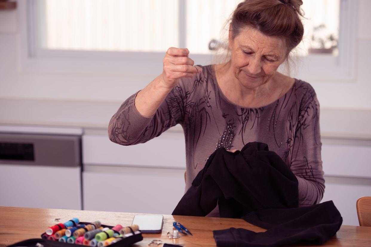 Senior woman darns her pants, sitting at the table.