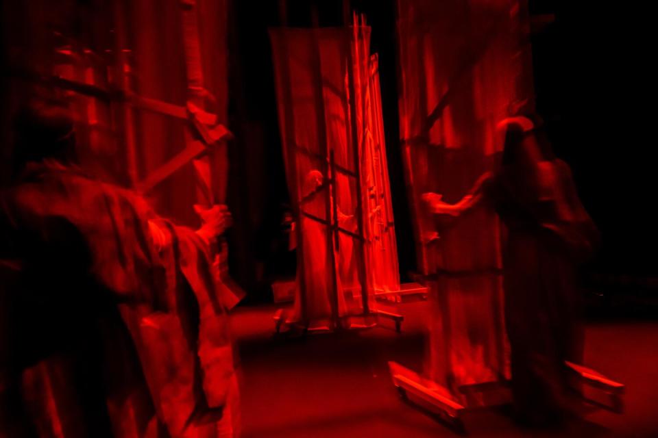 Actors bathed in blood-red light move set pieces of metal and gauzy white fabric.