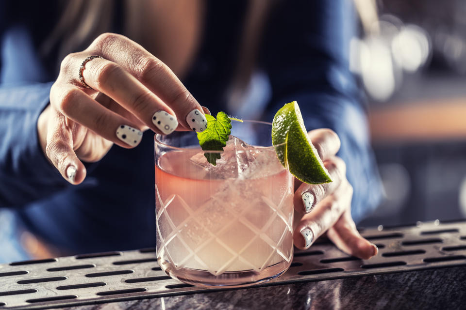 A person with polka dot nail art garnishes a cocktail with a lime wedge and mint leaf, placing it on a bar counter