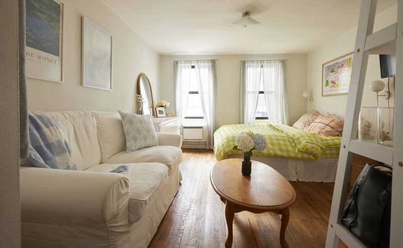 Living space with bed with chartreuse and white gingham duvet with floral and plaid pillowcases, straw round bedside table with case of yellow flowers, wood shoe rack with wall mount TV over it, windows with sheer white curtains, wood floor, White sofa with light blue and white pillows, oval wood table with vase of white and light blue hydrangeas