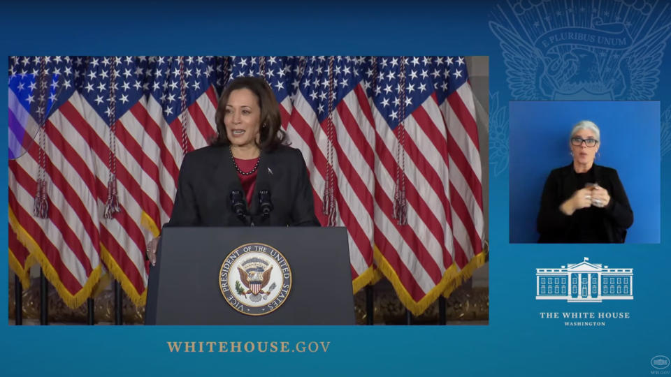 a woman in a dark suit speaks at a lectern with american flags in the background.