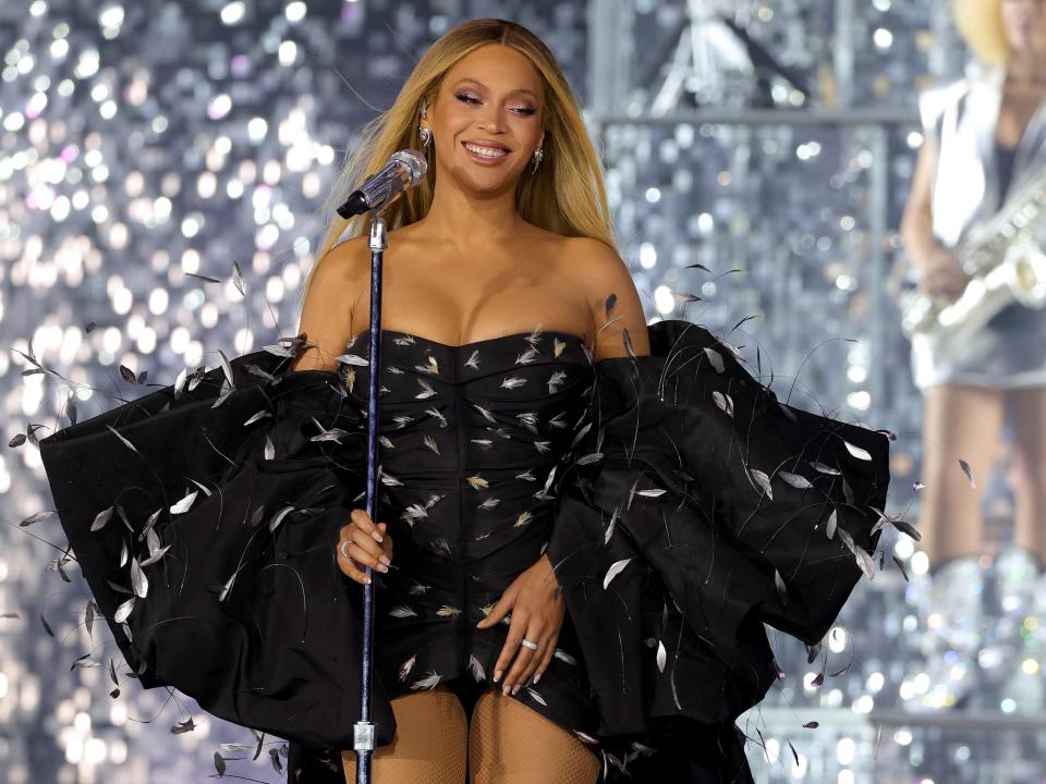 Beyonce smiling at her concert in Chicago while wearing a long black gown and stole with white feather details all over. She is holding the microphone stand with one hand