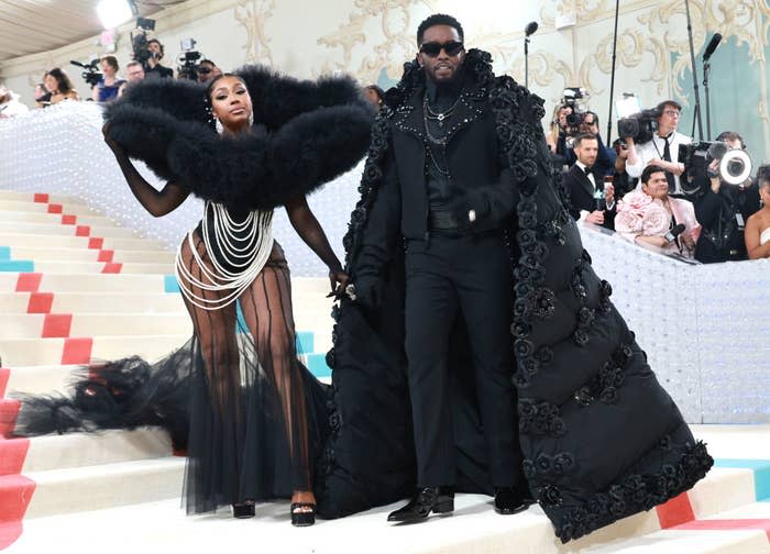 Yung Miami and Sean "Diddy" Combs on red carpet in dramatic black outfits with lavish feather and cape details at a celebrity event