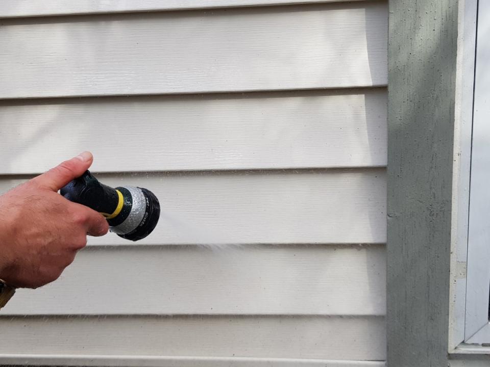 Person using a garden hose to spray grey vinyl siding on a house.