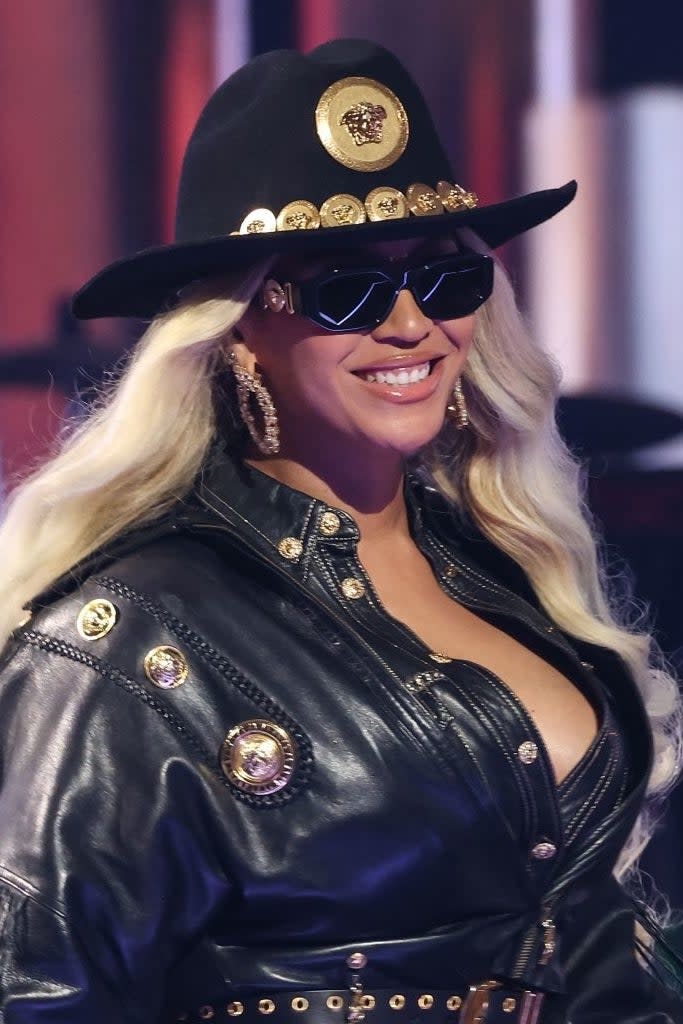 Beyoncé wears a studded black leather jacket and hat accessorized with gold embellishments and sunglasses, smiling on a stage