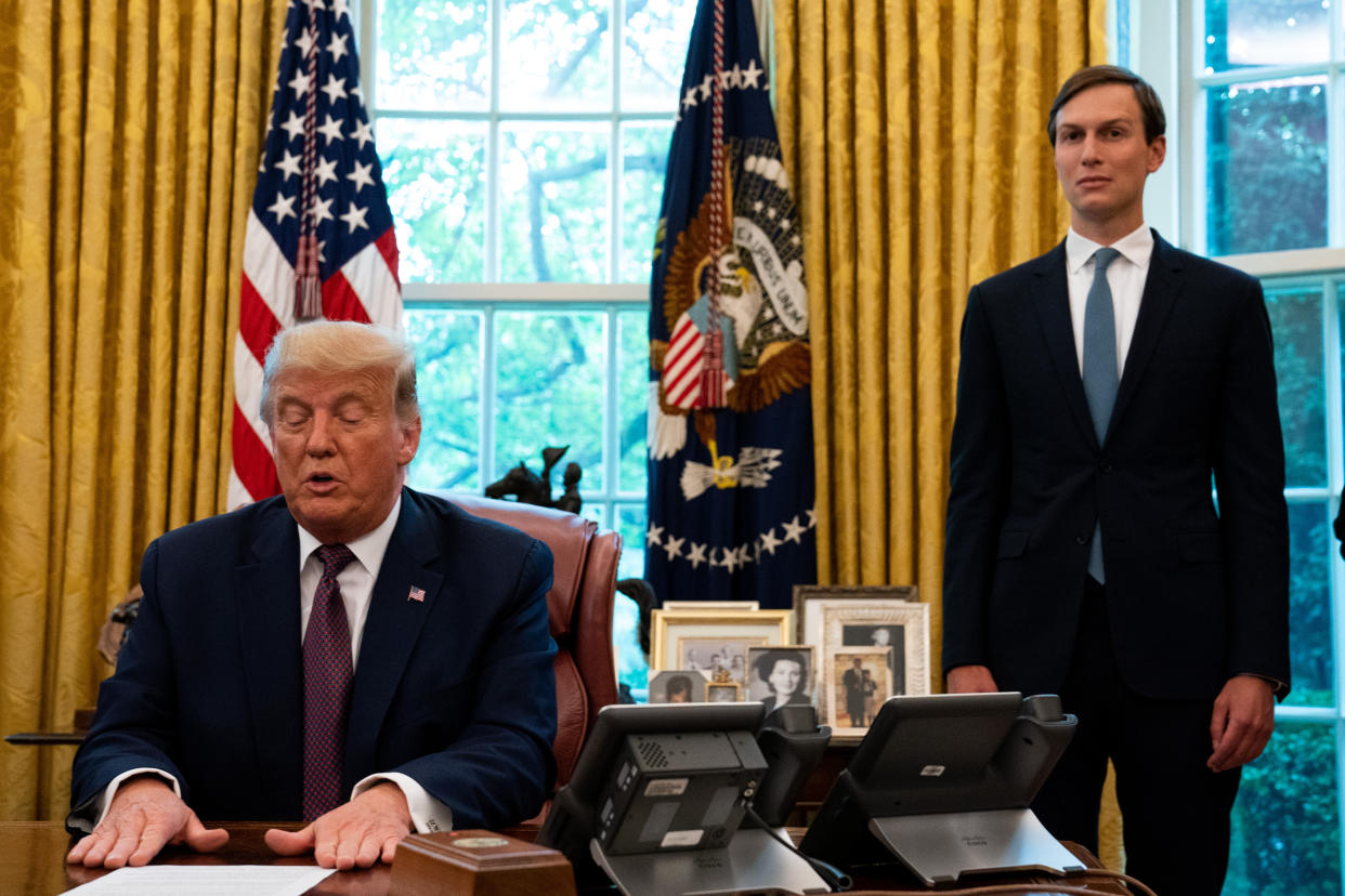 Jared Kushner stands stiffly next to two flags just off to the side of Donald Trump, who is seated at a desk.