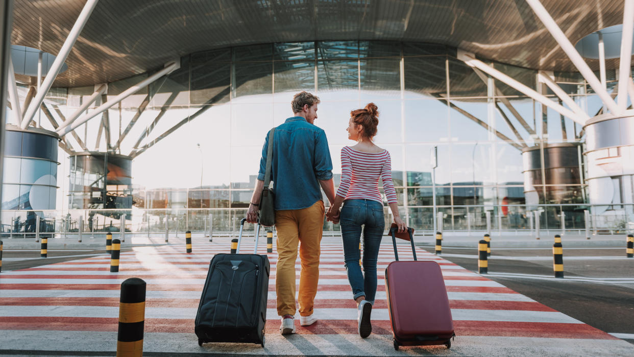 Full length back view portrait of young man and his charming girlfriend walking and carrying their trolley bags.