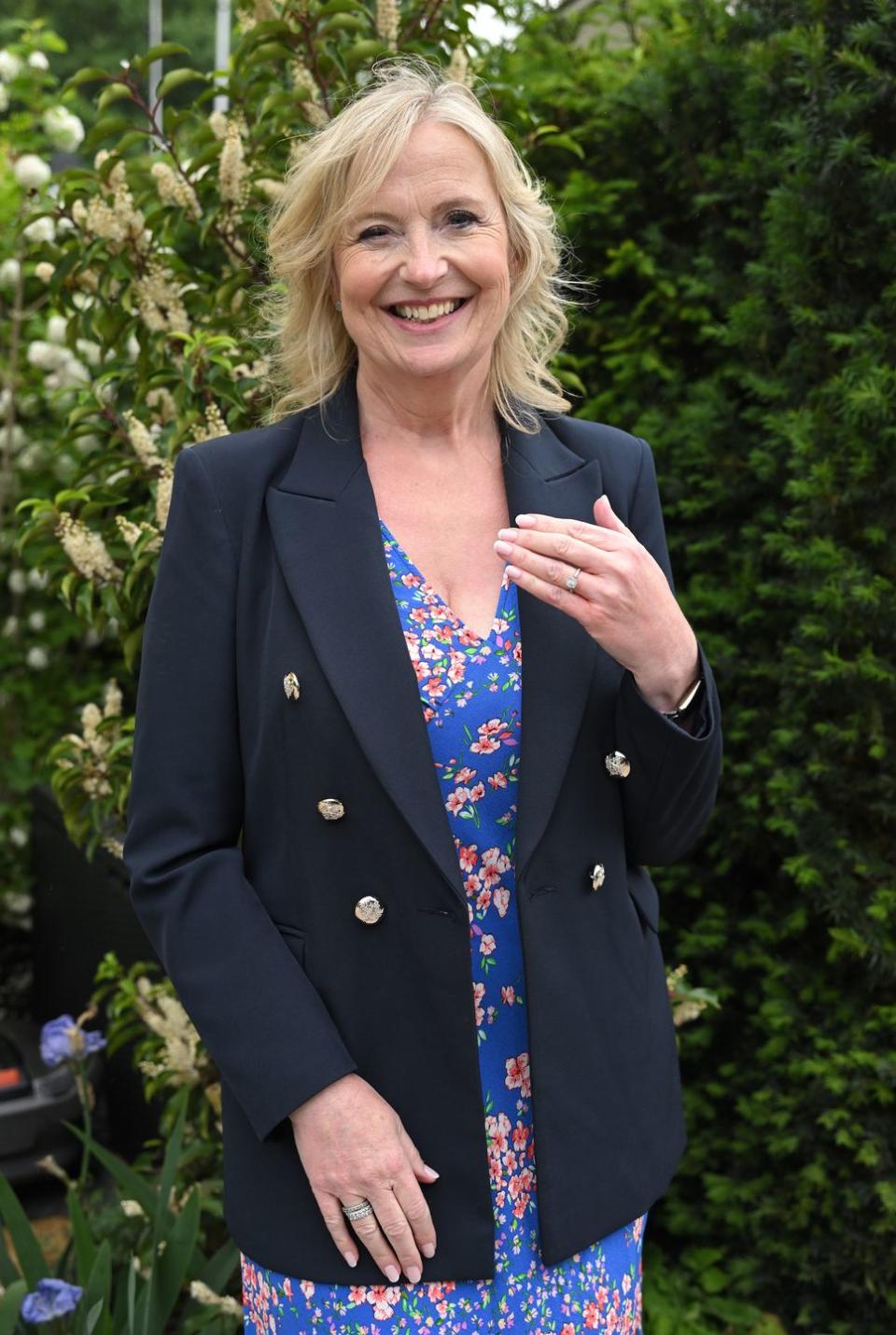 carol kirkwood smiles and holds up her hand to show photographers her engagement ring as she stands in front of a floral display