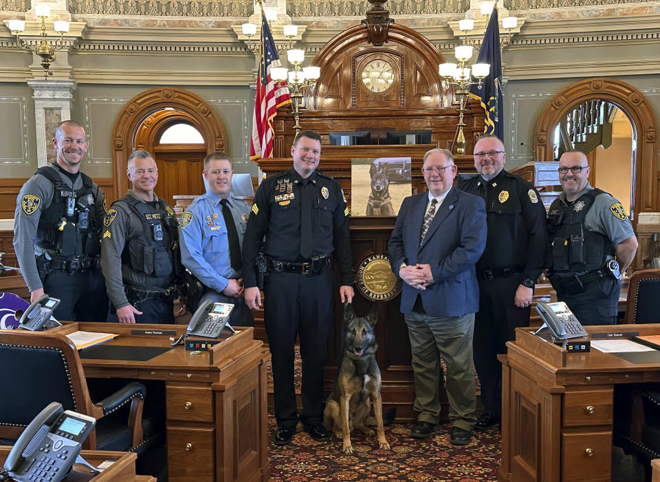 In this photo provided by the Kansas House of Representatives, Kansas House Speaker Dan Hawkins, R-Wichita, poses on Thursday, Feb. 1, 2024, with law enforcement officers from the Wichita area and Oz, a Wichita police dog, in the House chamber in Topeka, Kan. Oz's handler, Sedgwick County Sheriff's Deputy Tyler Brooks, appears left of the dog, while Hawkins appears to the dog's right. The photo behind the group is of Brooks' former K-9 partner, Bane, who was strangled to death by a domestic violence suspect. Brooks had a hand in drafting a bill to increase the penalties for killing police dogs. (Carrie Rahfaldt/Kansas House of Representatives via AP)