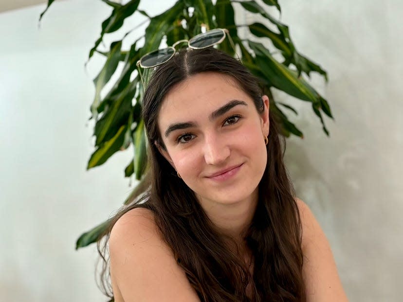 Sandrine Jacquot smiling and looking at the camera, she is sitting inside and has glasses on her head.  A plant is behind her.