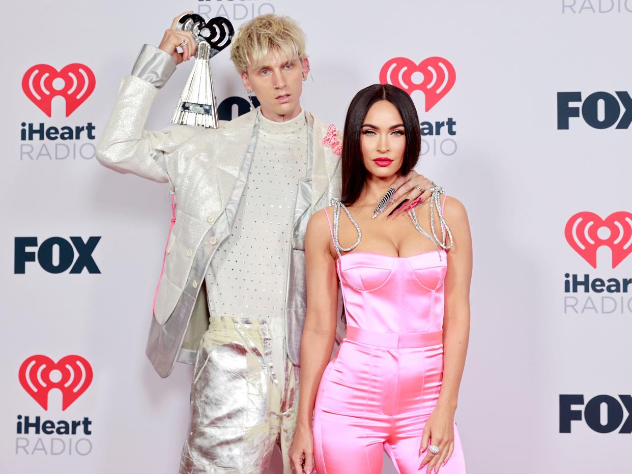 Rapper Machine Gun Kelly wears a white and silver suit and holds an iHeart Radio Music award while posing with his hand on girlfriend Megan Fox's neck, who is wearing a pink satin jumpsuit and staring at the camera.