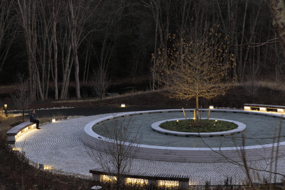 A visitor to the Sandy Hook Permanent Memorial sits on one of the benches during twilight, Monday, Dec. 5, 2022, in Newtown, Conn. The names of the 20 first graders and six educators killed a short distance away at Sandy Hook Elementary School 10 years ago are engraved in concrete around a memorial pool with a sycamore tree in the middle. (AP Photo/Julia Nikhinson)