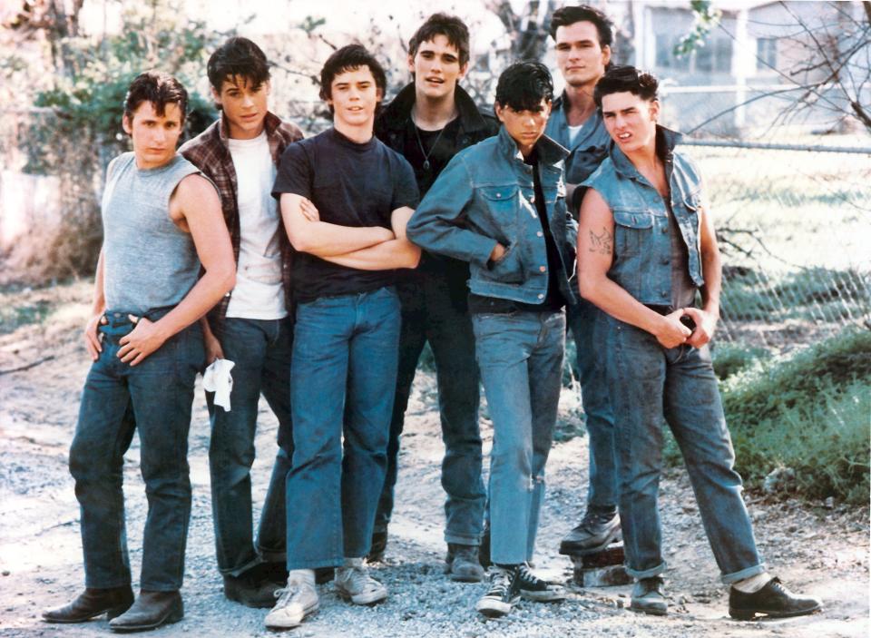 The cast of "The Outsiders" poses outdoors in casual 1980s attire: Patrick Swayze, Rob Lowe, Emilio Estevez, Tom Cruise, Matt Dillon, Ralph Macchio, and C. Thomas Howell