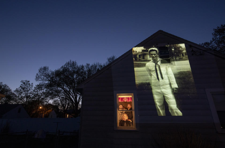 An image of veteran James Mandeville is projected onto the home of his daughter, Laurie Mandeville Beaudette, as she looks out a window with her son, Kyle, left, and husband, Mike, in Springfield, Mass., Tuesday, May 12, 2020. Mandeville, a U.S. Navy veteran and resident of the Soldier's Home in Holyoke, Mass., died from the COVID-19 virus at the age of 83. Seeking to capture moments of private mourning at a time of global isolation, the photographer used a projector to cast large images of veterans on to the homes as their loved ones are struggling to honor them during a lockdown that has sidelined many funeral traditions. (AP Photo/David Goldman)