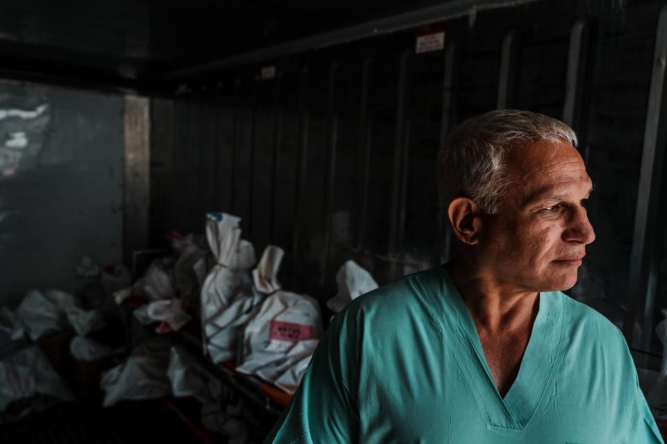 A man with graying hair, in blue scrubs, with numerous cinched white bags behind him