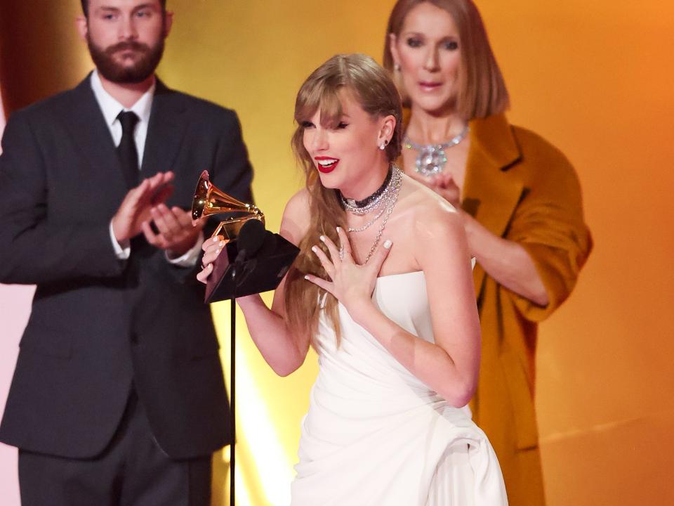 taylor swift onstage accepting a grammy award, holding the trophy in one hand and her other hand pressed to her chest. celine dion, in a brown overcoat, stands in the background