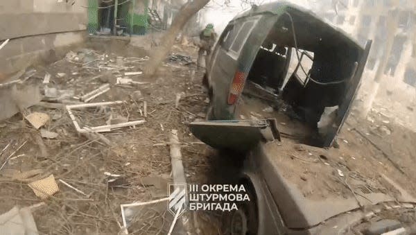 Footage of soldiers walking past a destroyed van, along a street strewn with debris.