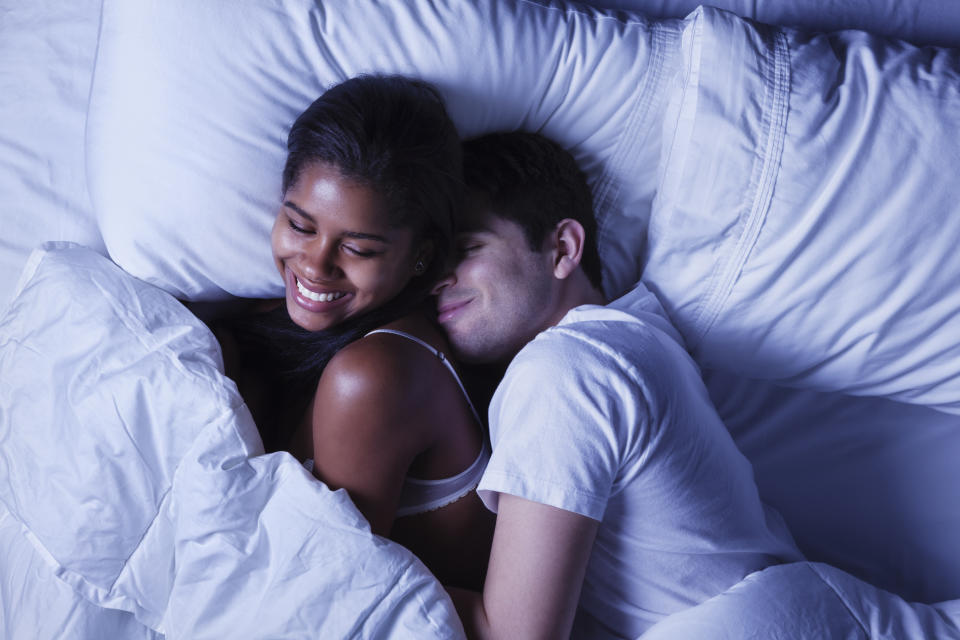 Photo of an attractive young mixed race couple, lying in bed as husband playfully invades his wife's side of the bed.