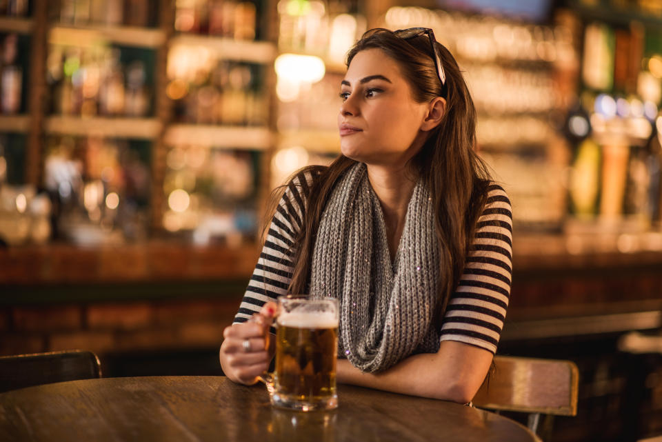 Beautiful pensive young woman holding a glass of beer while sitting in a pub and looking away.